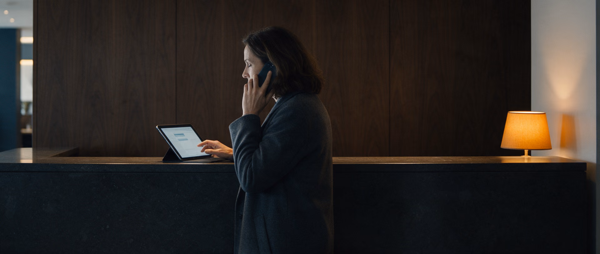 A woman stands at a dark stone ledge in a hotel lobby in the evening with a phone at her ear, a tablet in front of her and a lit lamp at the end of the ledge.