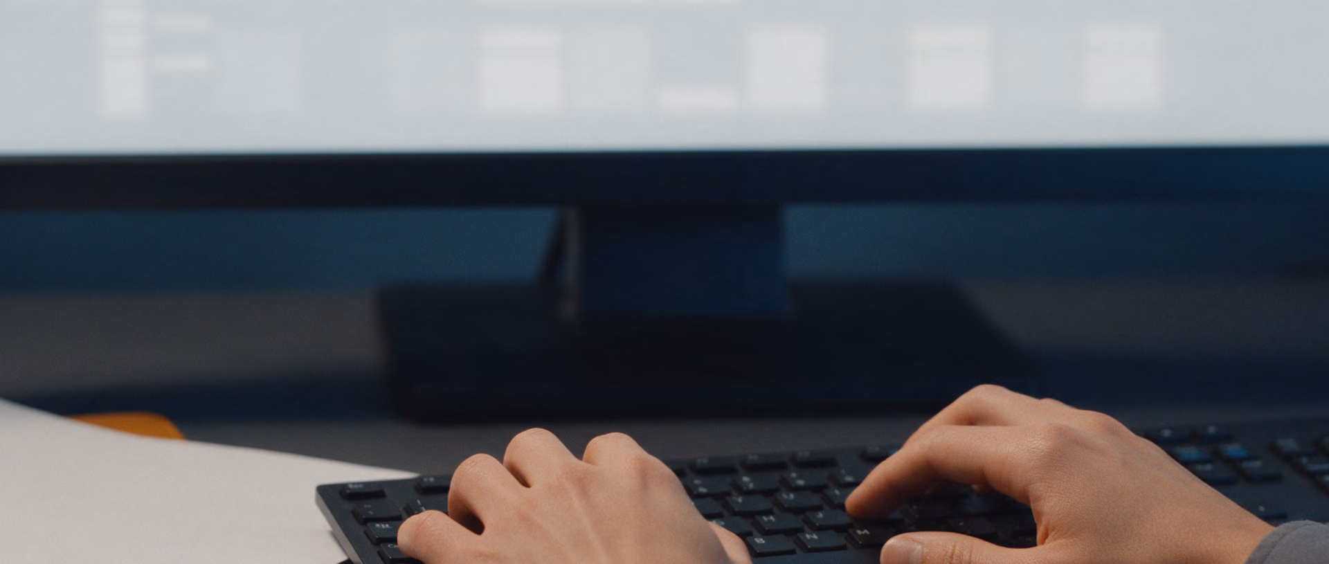Two hands on a keyboard in front of a monitor showing a blurred grid, with two blank sheets on a leather folder beside them.