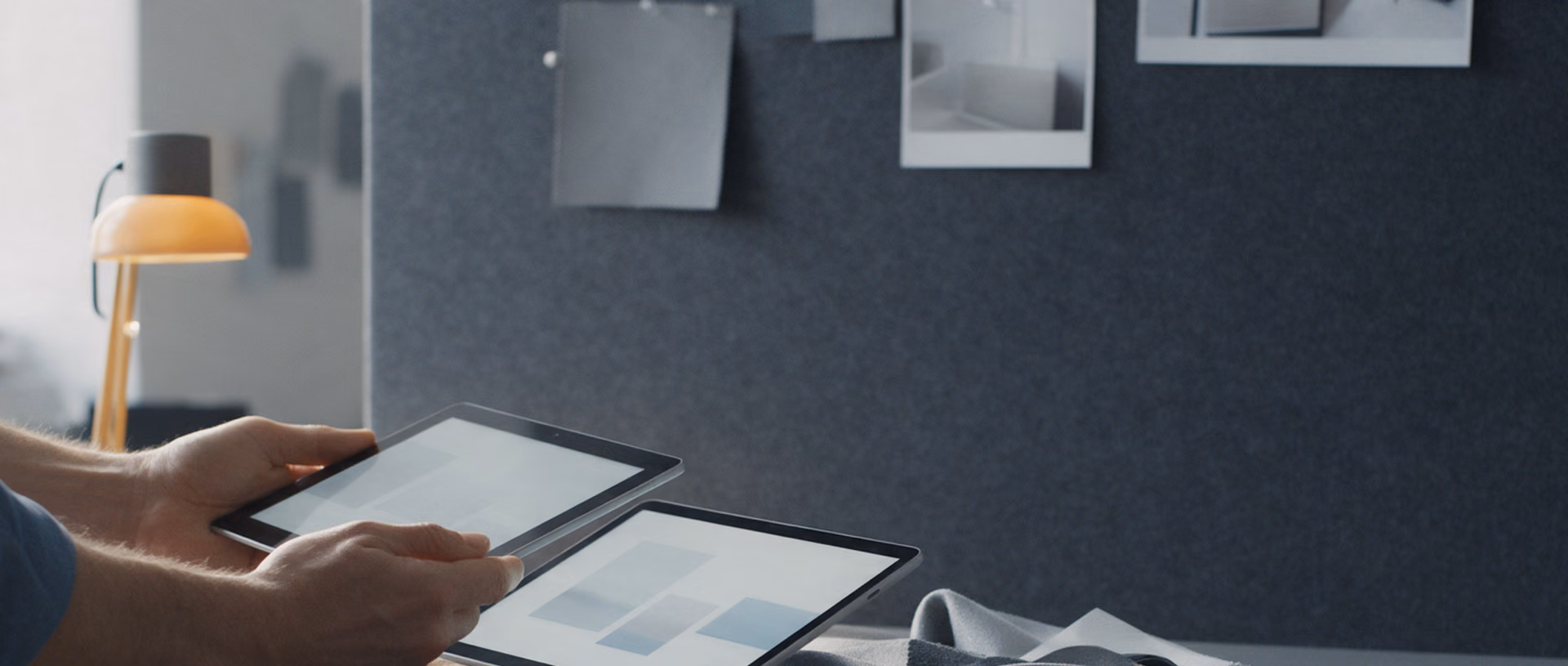 Two hands hold a tablet over a work table spread with fabric samples in front of a dark felt pinboard, a small desk lamp lit beside them.