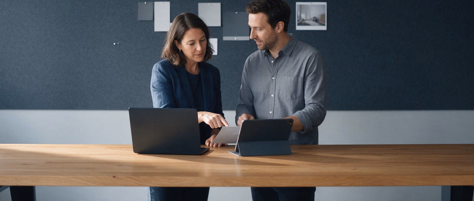 Two colleagues stand at a long oak table in front of a dark felt pinboard and compare a laptop and a tablet whose backs face the camera.