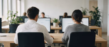 Two colleagues seen from behind, working side by side at their monitors in a bright office with green plants.