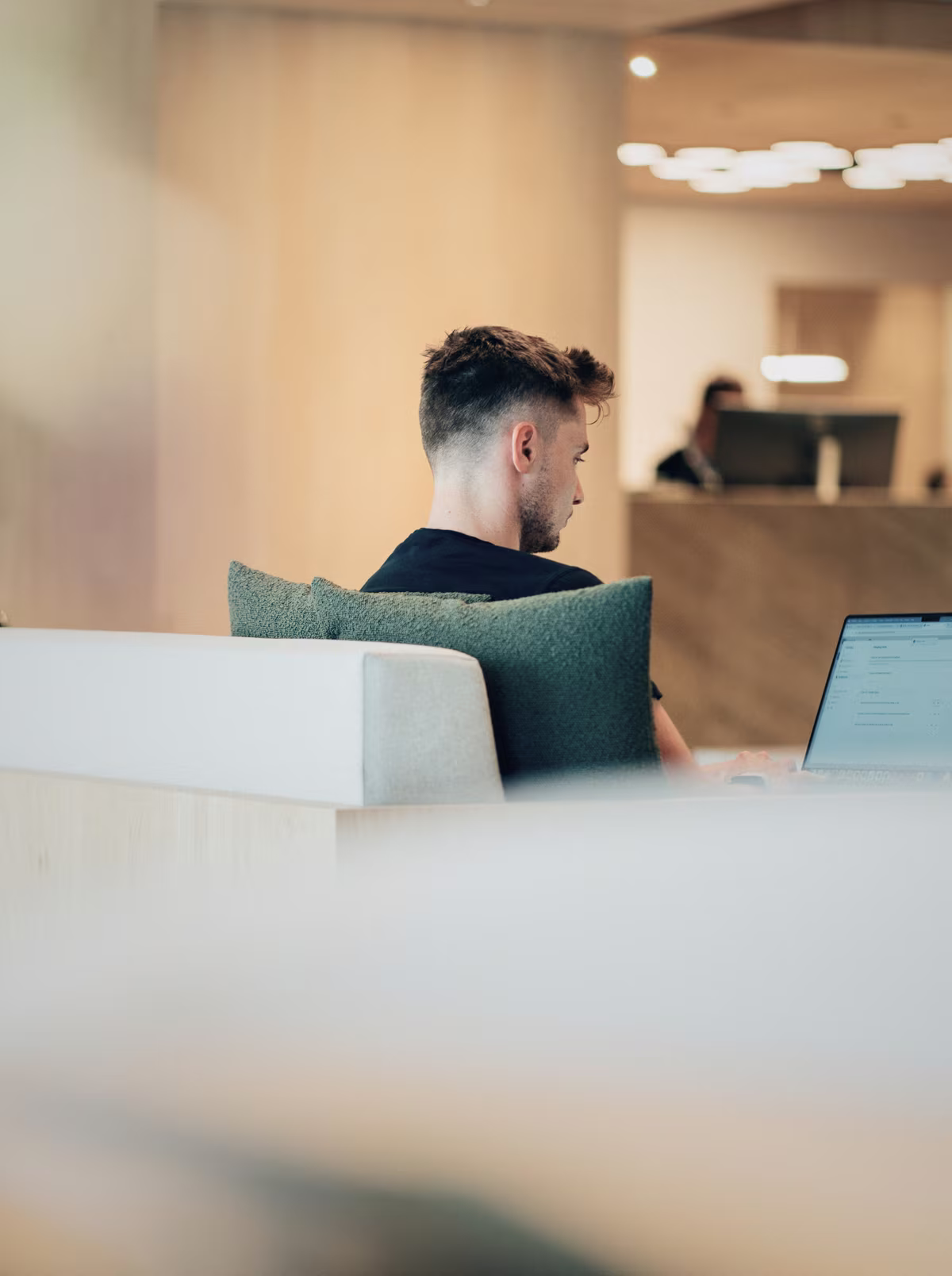 A colleague working at a laptop in the lounge, with a second colleague at the reception desk behind.